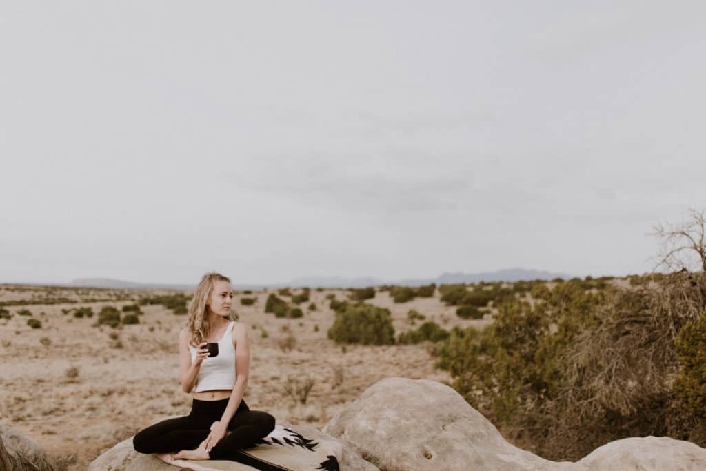 Girl Holding Coffee while Sitting on Southwest Style Blanket and does Yoga in front of a Desert Landscape View in Santa Fe New Mexico